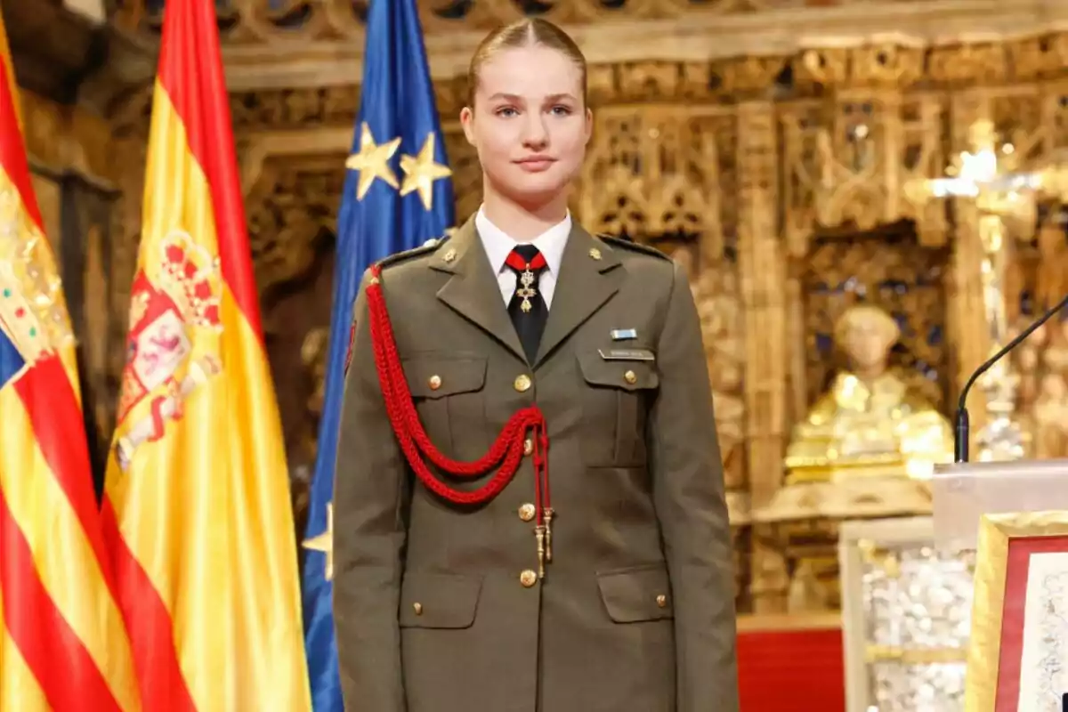 Una joven vestida con uniforme militar se encuentra de pie frente a las banderas de España y la Unión Europea en un entorno decorado con detalles dorados.