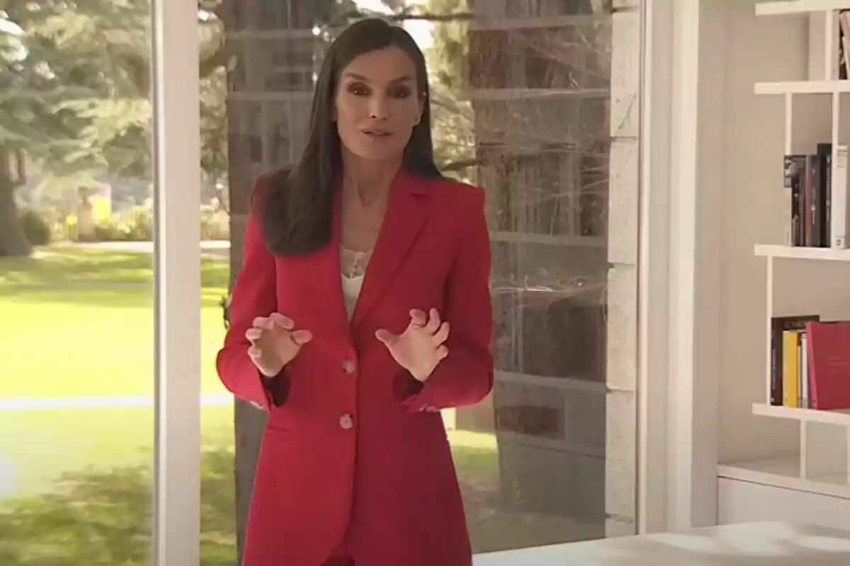 Una mujer con traje rojo está de pie en una habitación luminosa con una ventana grande y estanterías con libros.