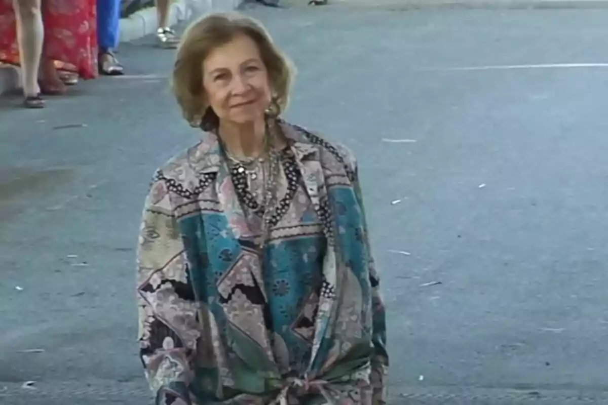 Una mujer mayor con cabello corto y canoso, vestida con una chaqueta de colores y patrones variados, está de pie en una calle pavimentada mientras otras personas se ven al fondo.