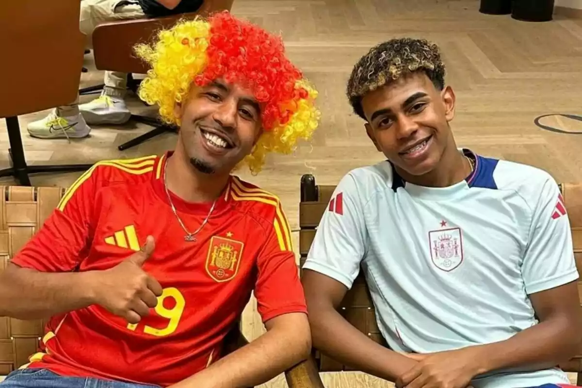 Dos personas sonrientes con camisetas de la selección española de fútbol, una de ellas lleva una peluca de colores rojo y amarillo.