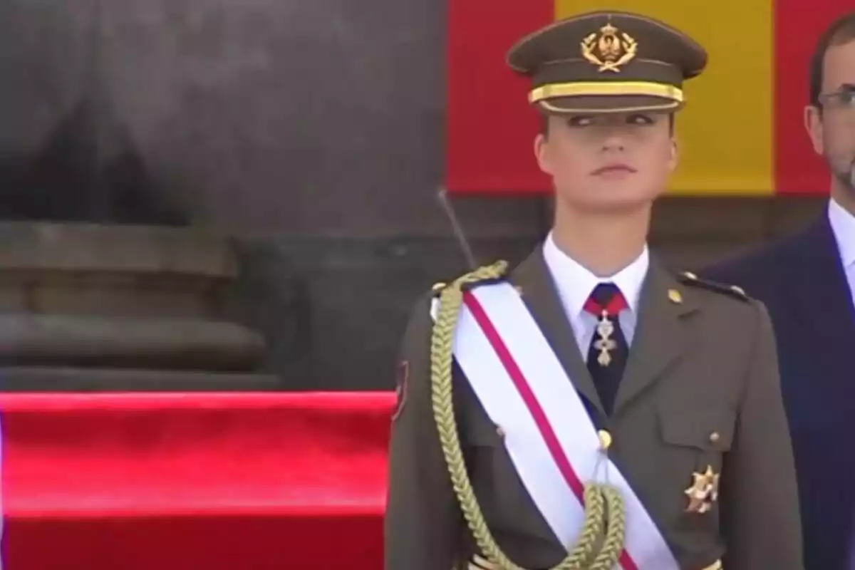 Una mujer en uniforme militar con una banda blanca y roja, de pie frente a una bandera.