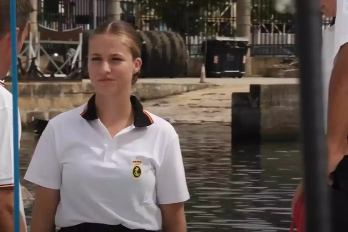 Una joven con una camiseta blanca y un escudo en el pecho está de pie cerca de un muelle, con el agua y algunas estructuras en el fondo.