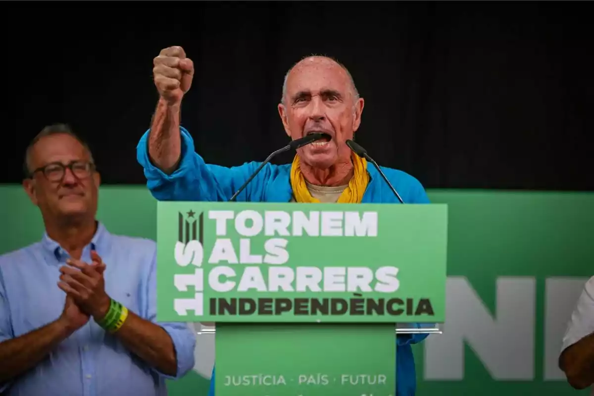 Hombre hablando en un mitin con un cartel verde que dice "Tornem als carrers independència" y otra persona aplaudiendo al fondo.