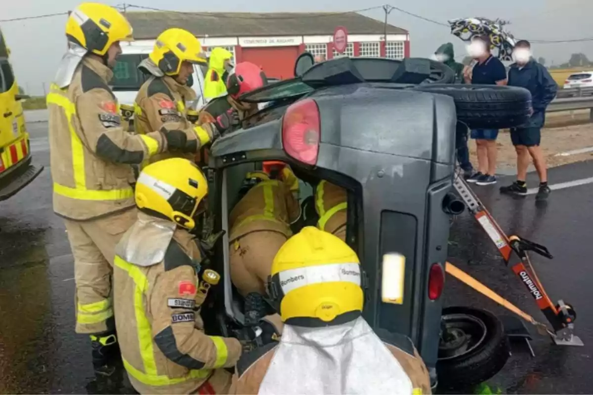 Bomberos trabajando en el rescate de personas atrapadas en un coche volcado en la carretera mientras algunos transeúntes observan.