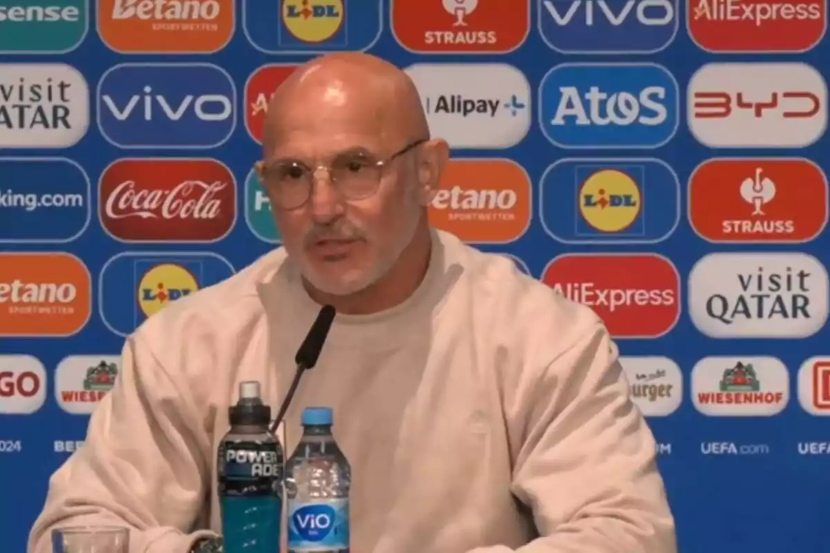 Un hombre calvo con gafas y barba corta, vestido con un suéter beige, habla en una conferencia de prensa con un micrófono frente a él y varias botellas de agua y bebidas energéticas sobre la mesa, con un fondo lleno de logotipos de patrocinadores.