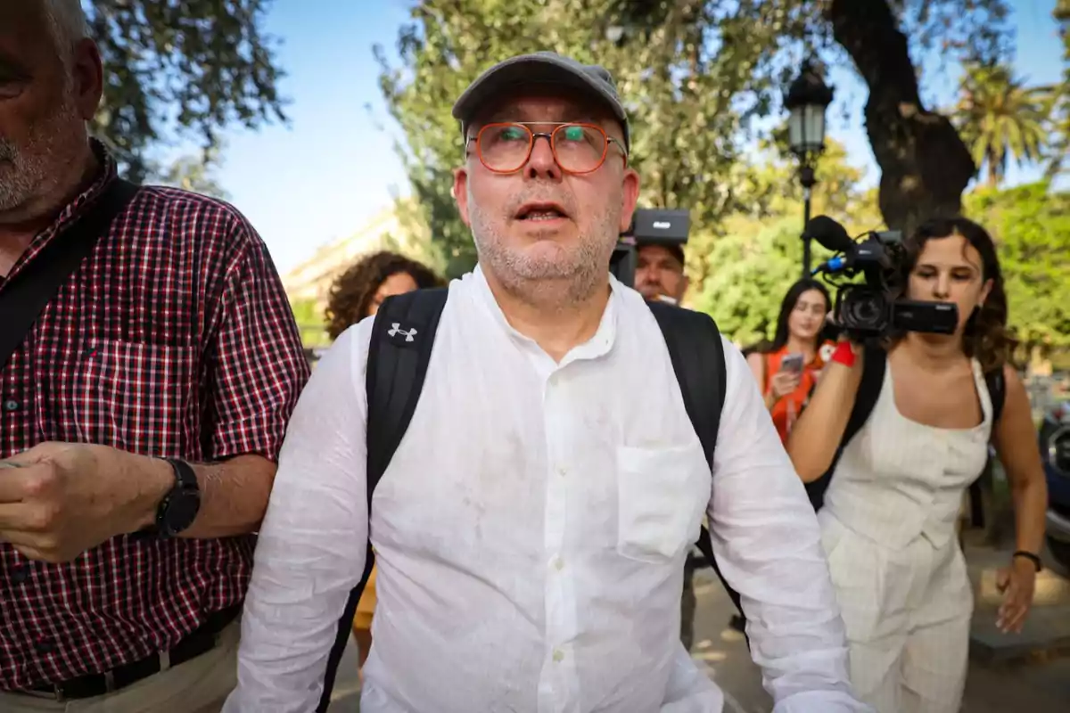 Un hombre con gafas y gorra, vestido con una camisa blanca y mochila, rodeado de personas y cámaras en un entorno al aire libre con árboles.