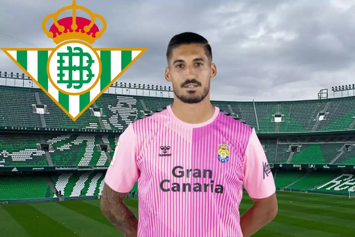 Un jugador de fútbol con una camiseta rosa de Gran Canaria está de pie en un estadio de fútbol con el logo del Real Betis en el fondo.