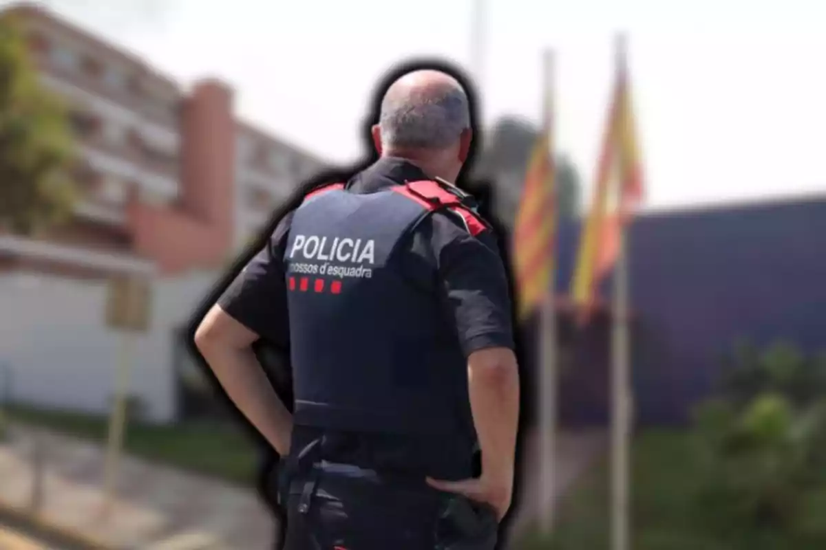 Un agente de policía de espaldas con uniforme de los Mossos d'Esquadra en un entorno urbano con banderas al fondo.