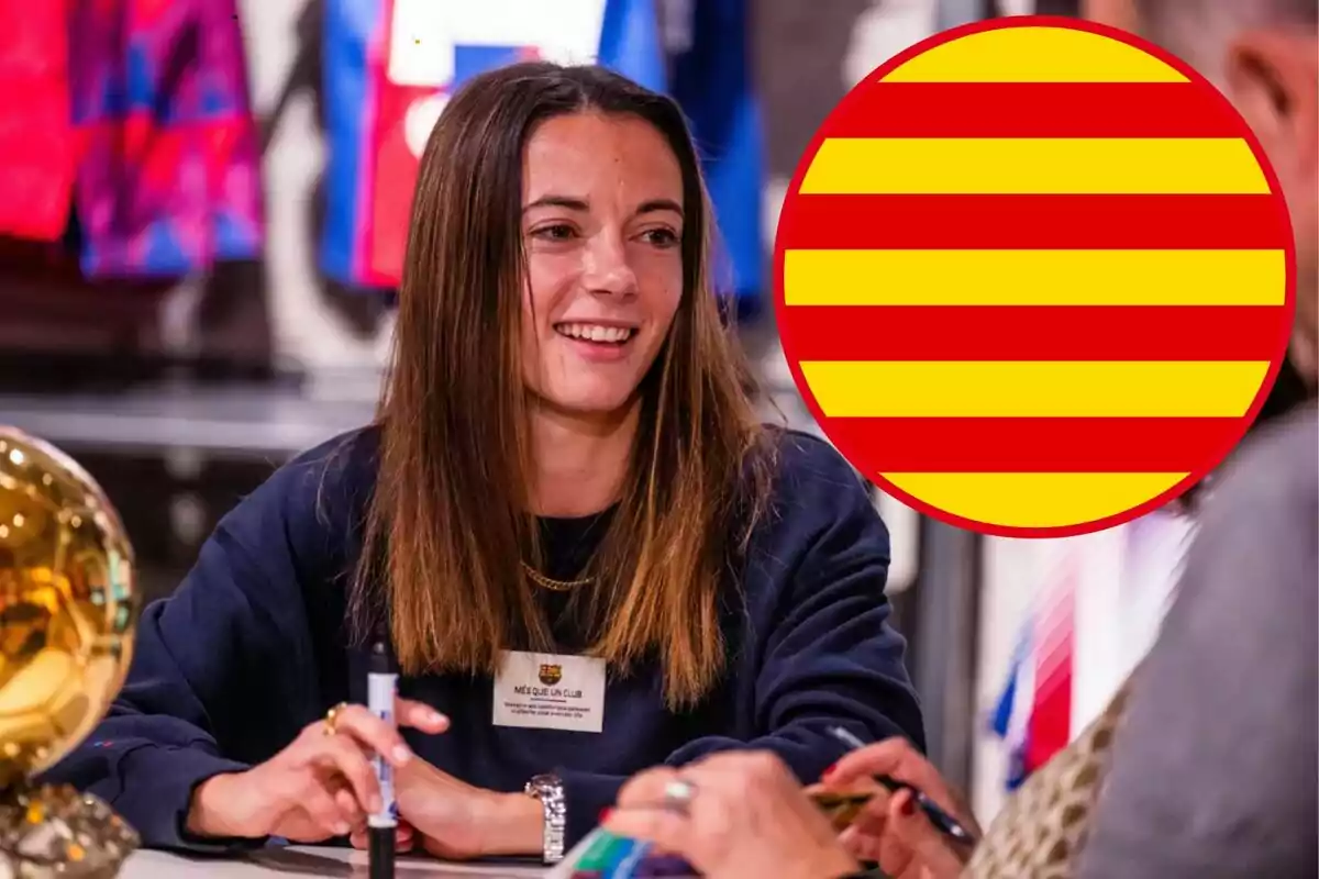 Una mujer sonriente con cabello largo y lacio, sentada en una mesa firmando autógrafos, con una pelota dorada a su lado y una insignia de rayas rojas y amarillas en la esquina derecha de la imagen.