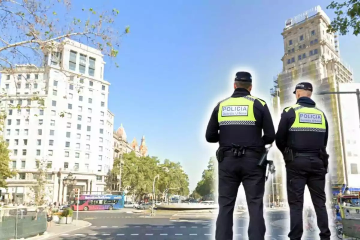 Dos agentes de la Guardia Urbana de espaldas observan una intersección en una ciudad con edificios altos y un autobús de colores en el fondo.