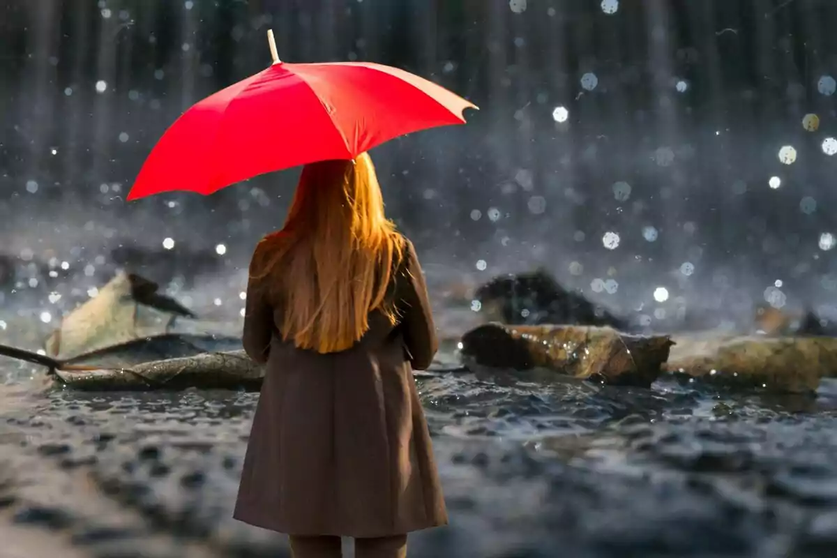 Persona con cabello largo y abrigo marrón sosteniendo un paraguas rojo bajo la lluvia.