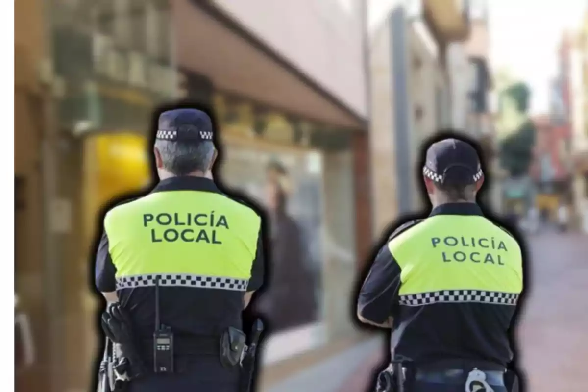 Dos agentes de policía local de espaldas con chalecos reflectantes en una calle urbana.