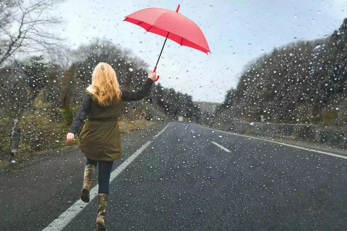 Persona caminando bajo la lluvia en una carretera con un paraguas rojo.