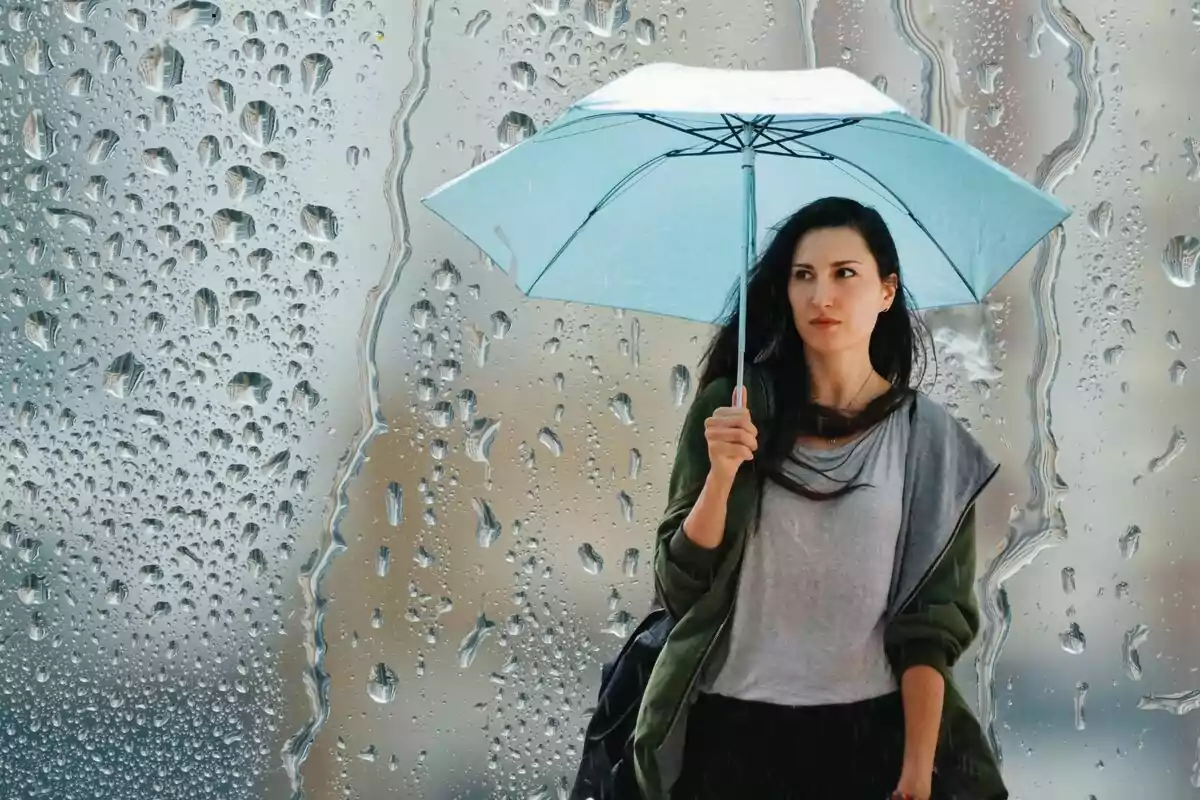 Una mujer con una sombrilla azul camina bajo la lluvia, con gotas de agua en una ventana en el fondo.