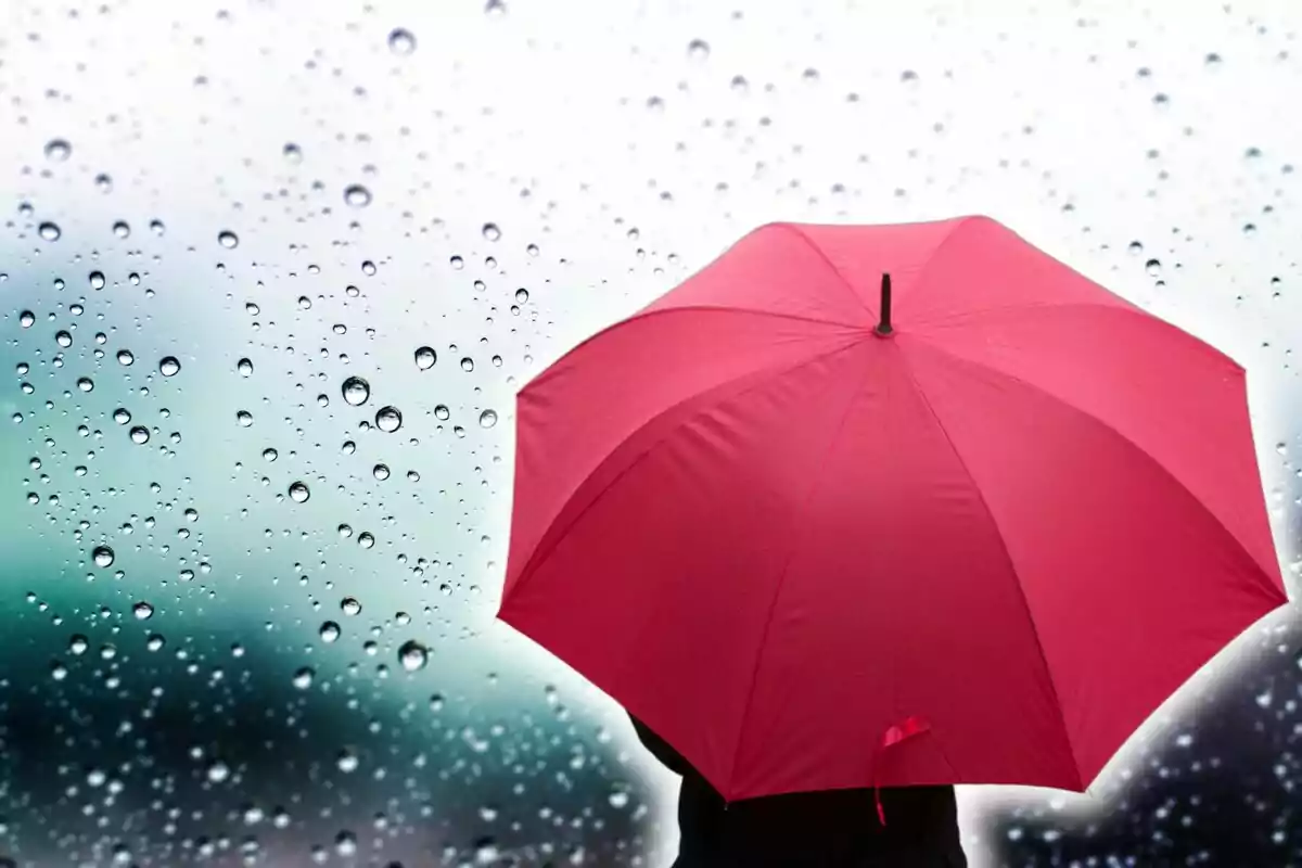 Persona sosteniendo un paraguas rojo bajo la lluvia con gotas de agua en una ventana.