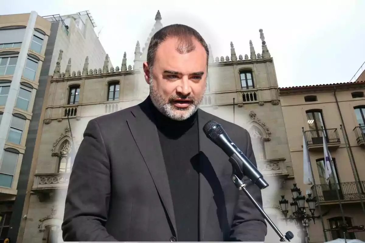 Un hombre con barba y cabello corto habla frente a un micrófono con un edificio histórico de fondo.