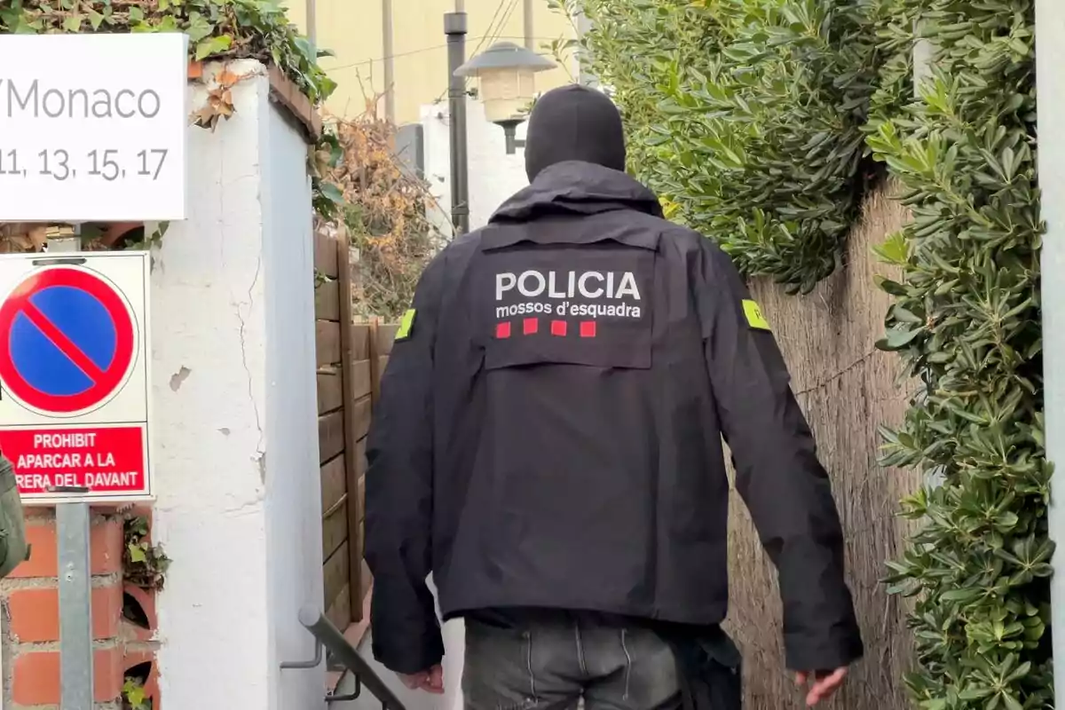 Un agente de la policía de los Mossos d'Esquadra camina por un pasillo exterior rodeado de vegetación, junto a un cartel que indica la prohibición de aparcar.