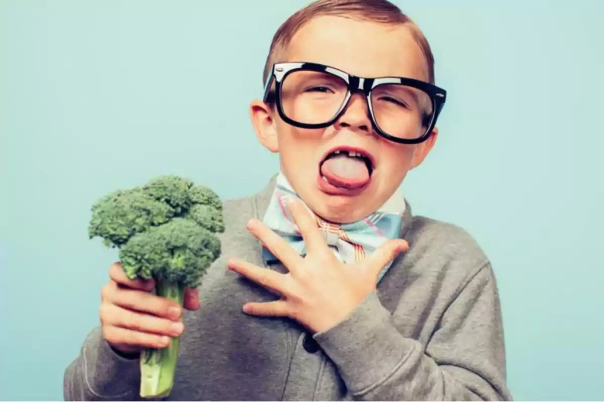 Niño con gafas grandes haciendo una mueca mientras sostiene un brócoli.