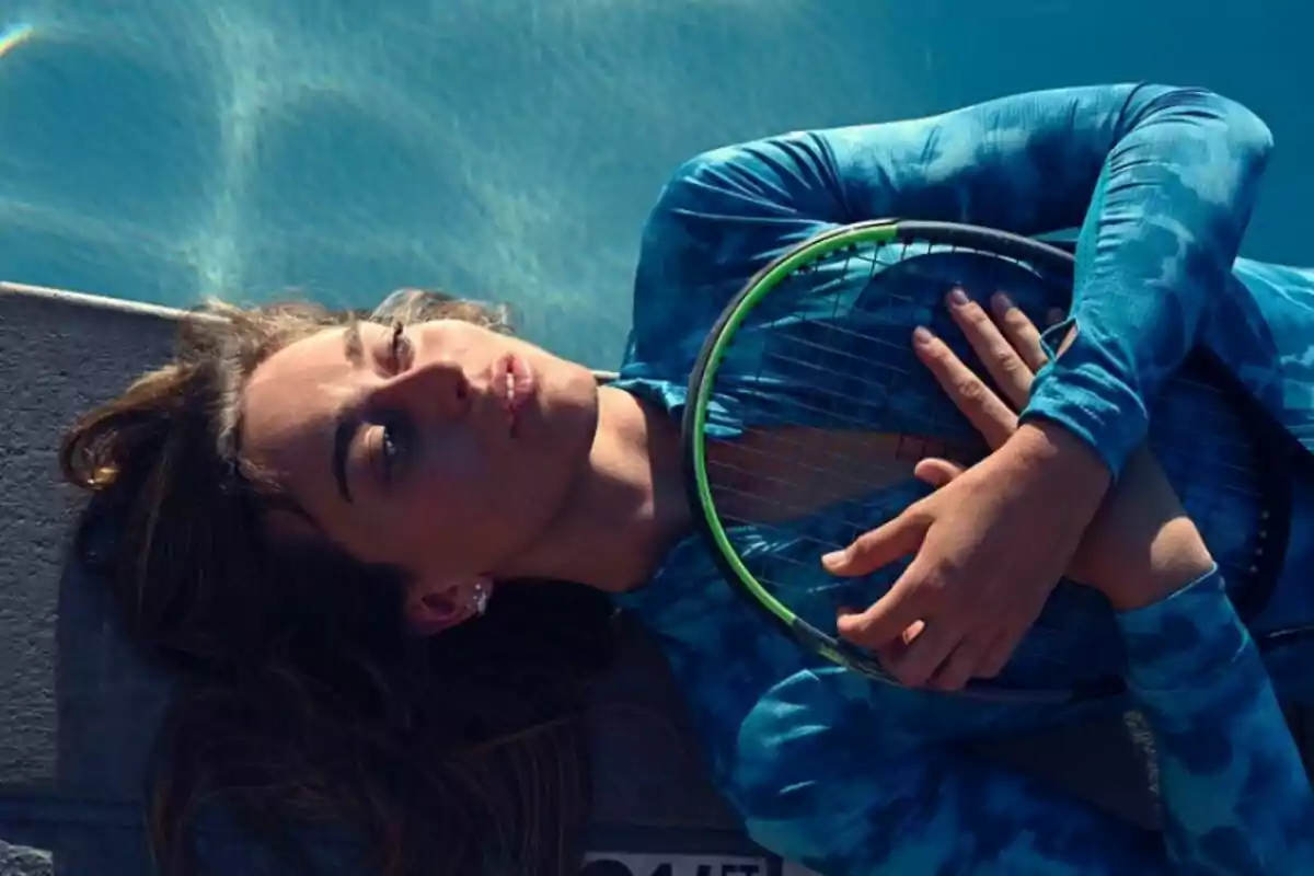 Una persona recostada junto a una piscina, sosteniendo una raqueta de tenis sobre su pecho y vistiendo una camiseta de manga larga azul.