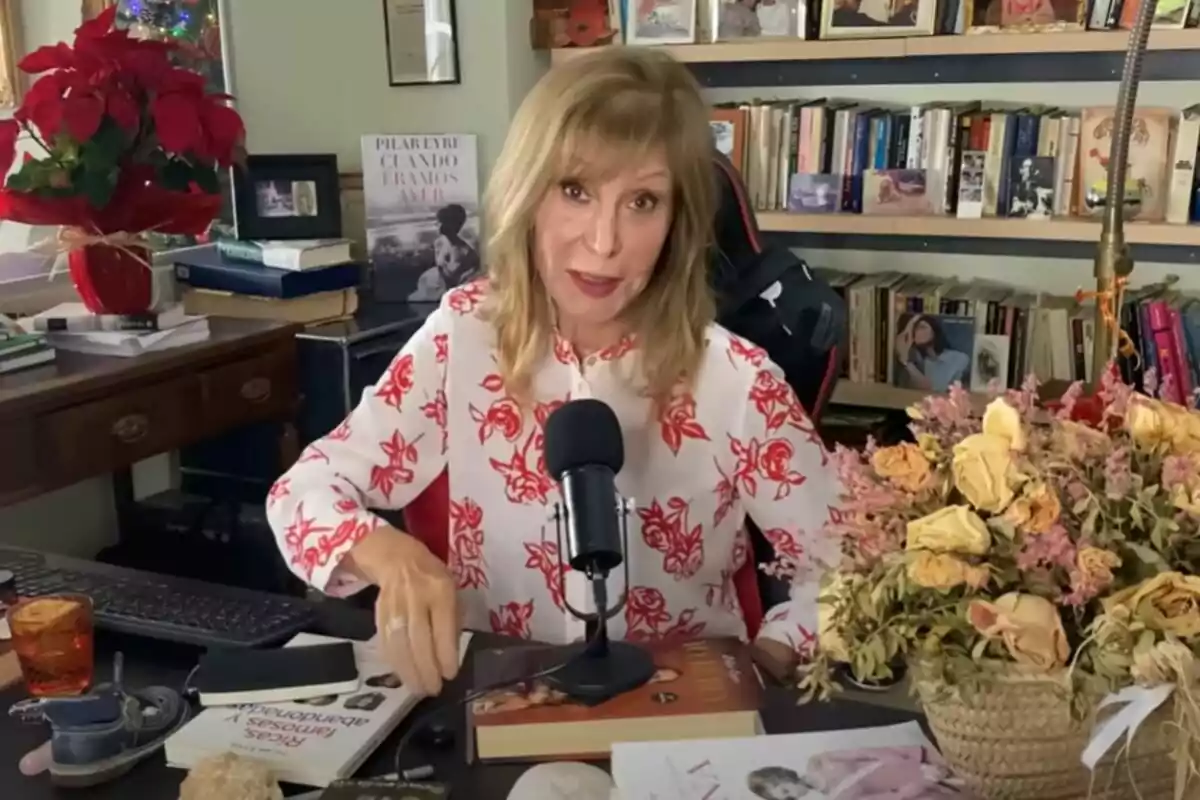 Una mujer de cabello rubio y blusa blanca con flores rojas está sentada frente a un micrófono en una habitación llena de libros y decoraciones, incluyendo una planta de poinsettia roja y un ramo de flores secas.