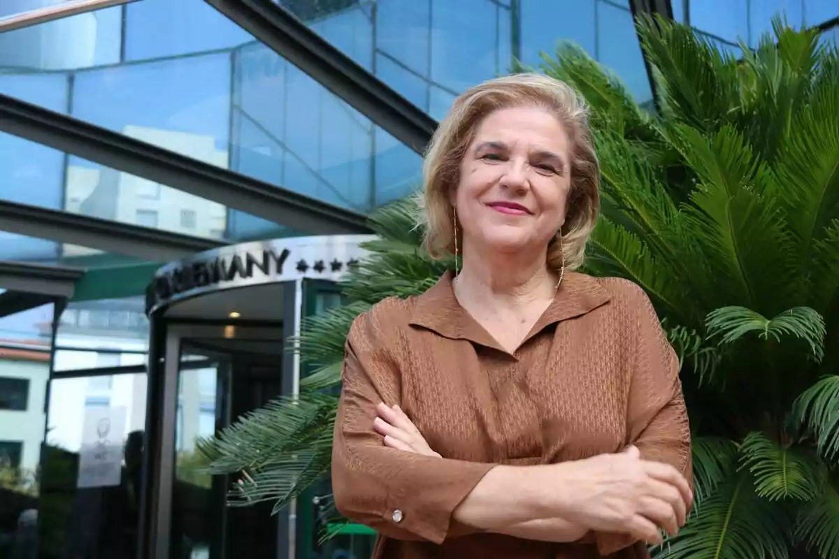 Mujer sonriendo con los brazos cruzados frente a un edificio de cristal y plantas verdes.