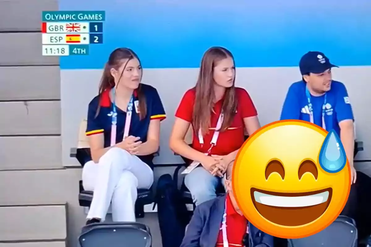 Tres personas sentadas en las gradas durante un partido de los Juegos Olímpicos, con el marcador mostrando que España está ganando 2-1 a Gran Bretaña en el cuarto periodo, y un emoji grande cubriendo parcialmente la imagen.