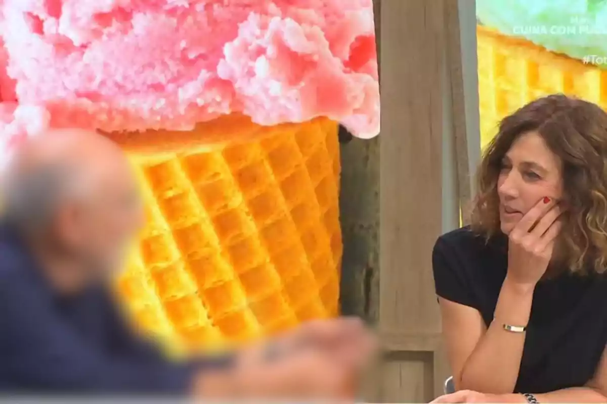 Una mujer con cabello rizado y una camiseta negra está sentada frente a un hombre desenfocado, con un fondo de un gran helado de fresa en un cono.