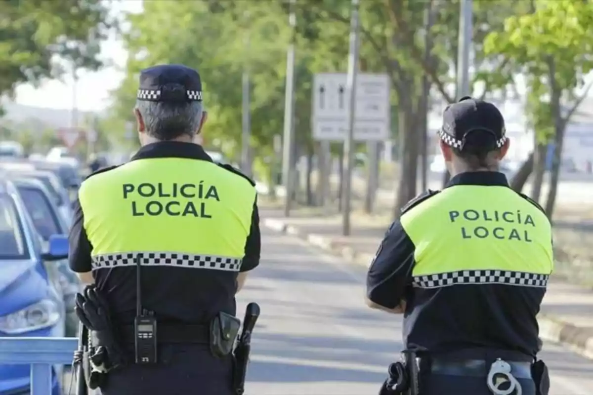 Dos agentes de la policía local de espaldas con chalecos reflectantes amarillos y uniformes negros patrullando una calle.