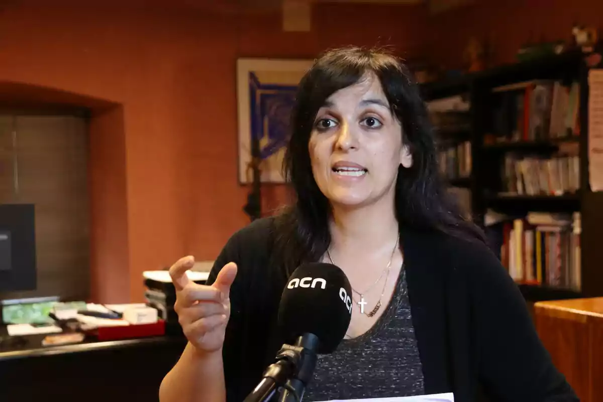 Mujer hablando frente a un micrófono en una habitación con estanterías llenas de libros.