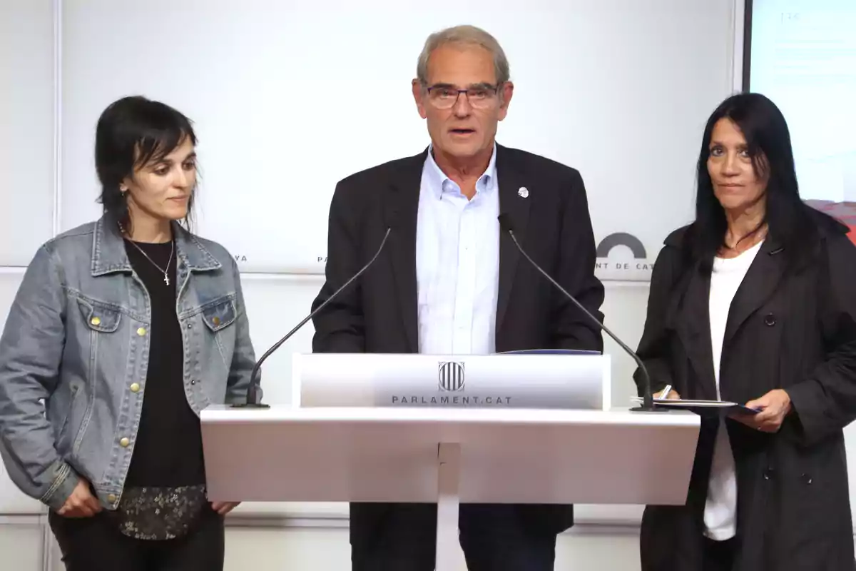 Tres personas de pie en un podio con el logotipo del Parlamento de Cataluña, una mujer con chaqueta de mezclilla a la izquierda, un hombre con gafas y traje en el centro, y una mujer con chaqueta negra a la derecha.