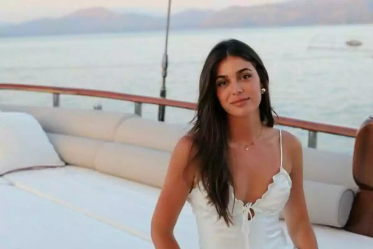 Mujer con vestido blanco sentada en un yate con el mar y montañas de fondo.