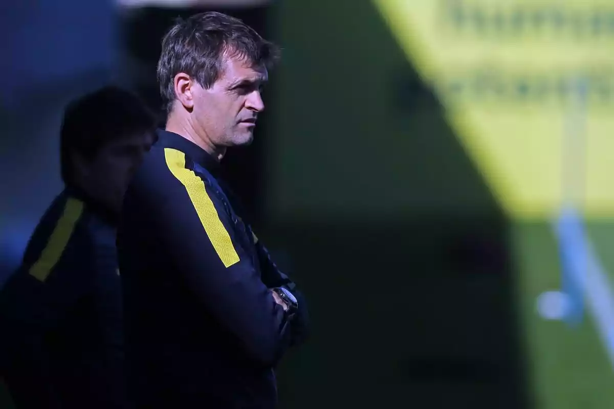 Un hombre con uniforme deportivo negro y amarillo está de pie con los brazos cruzados, mirando hacia la derecha en un entorno con luz y sombra.