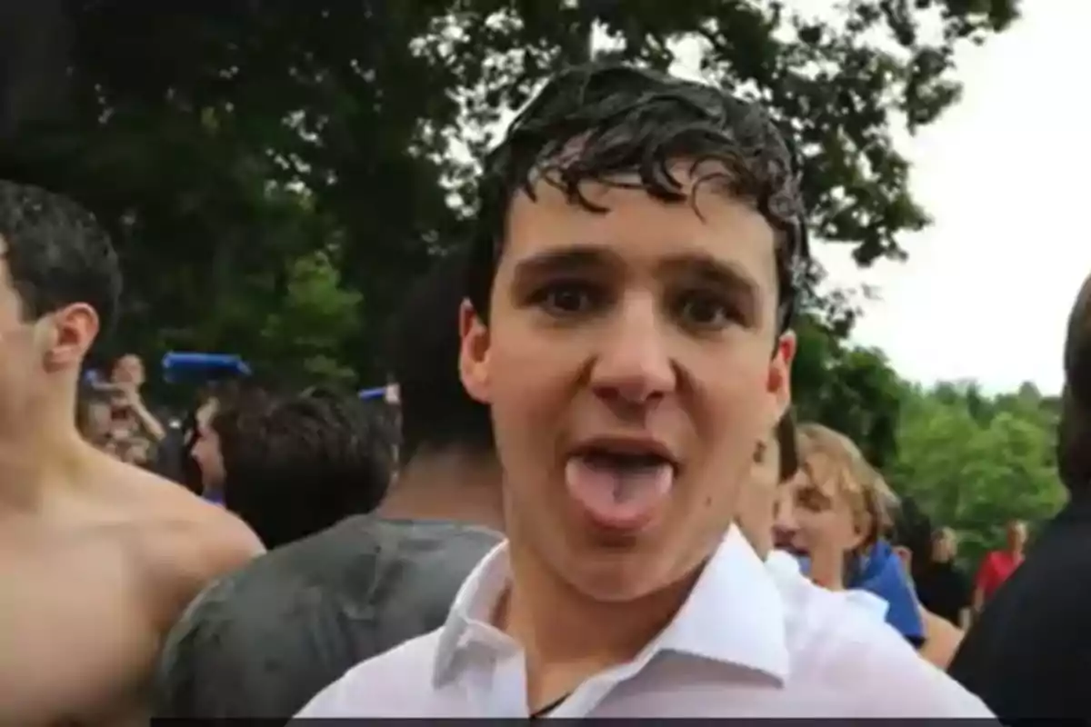 Un joven con la camisa mojada saca la lengua mientras está rodeado de otras personas en un entorno al aire libre.