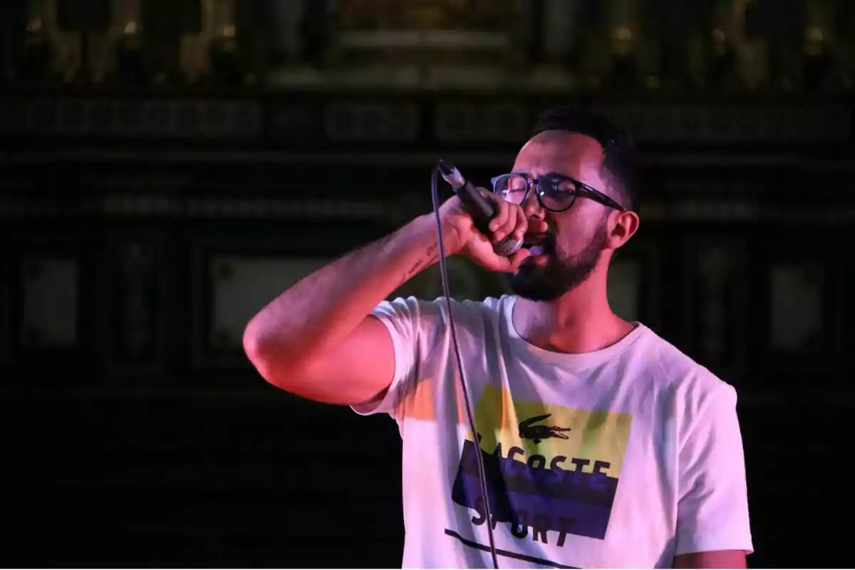 Un hombre con gafas y barba canta en un micrófono mientras usa una camiseta blanca con el logo de Lacoste Sport.