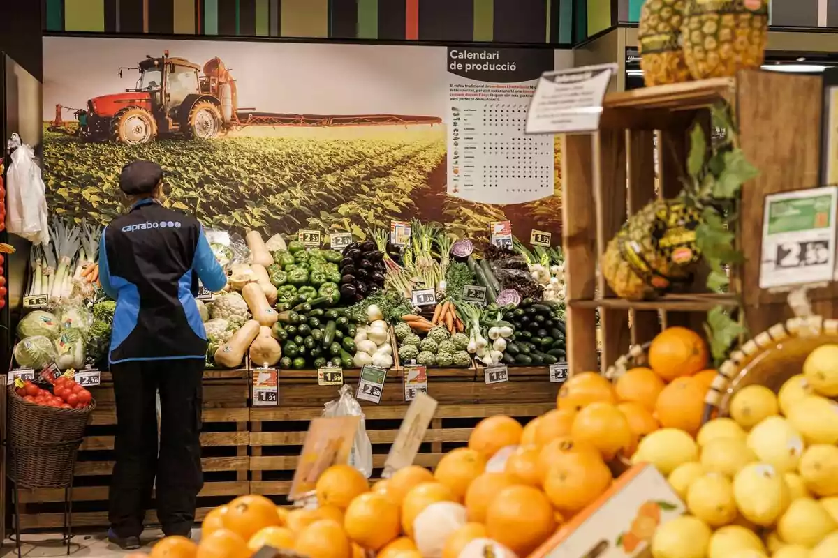 Un empleado de supermercado organiza verduras y frutas frescas en una sección de productos agrícolas, con un fondo que muestra un campo de cultivo y un calendario de producción.