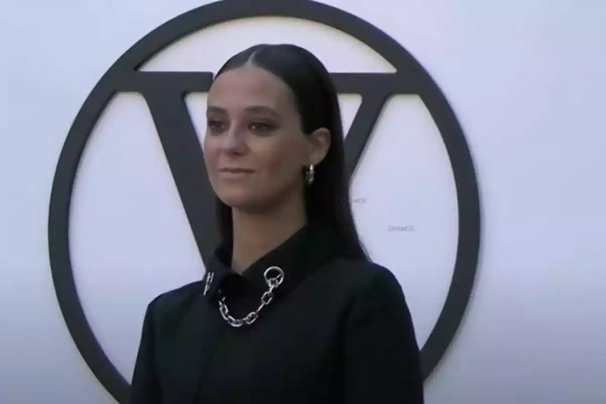 Una mujer con cabello oscuro y lacio, vestida de negro, posa frente a un logotipo circular con una letra 